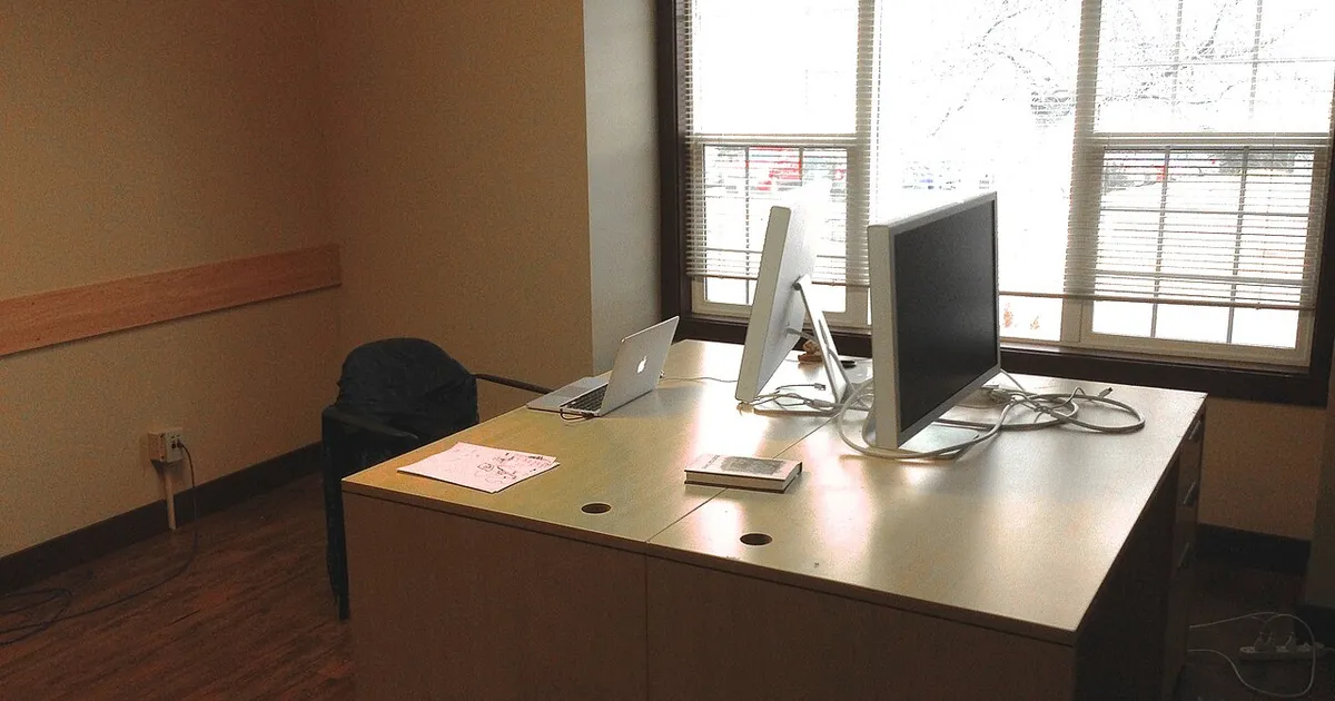 A bright room with a corner desk, two monitors and a laptop beside a large window